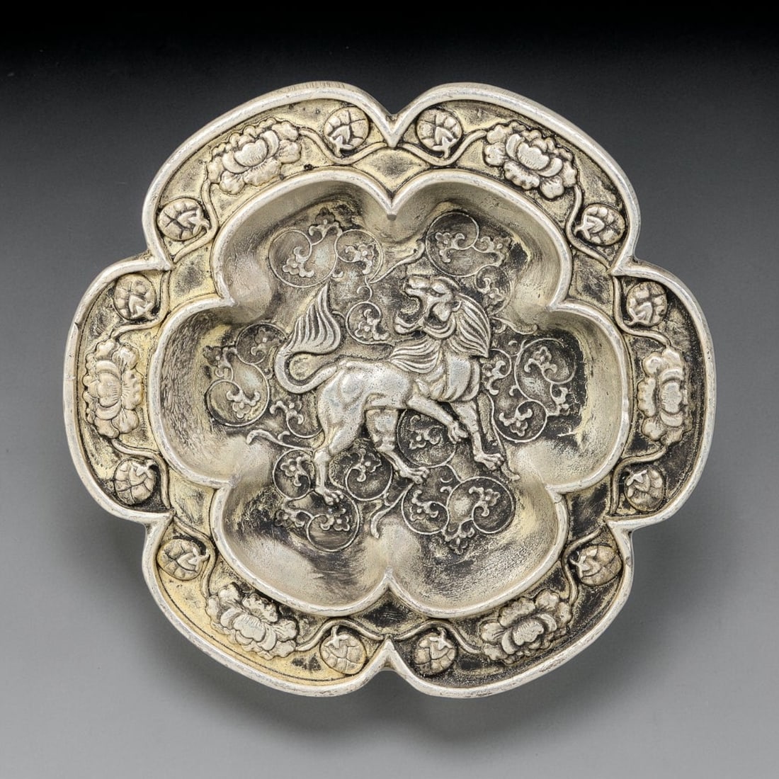 A Chinese German Silver Lion Lobed Dish, 48BD: Cast German silver lobed dish with a quatrefoil form. The interior face is decorated in relief with a lion amid scrolling foliate motifs. The raised rim bears repeating floral and seed pod ornamentati