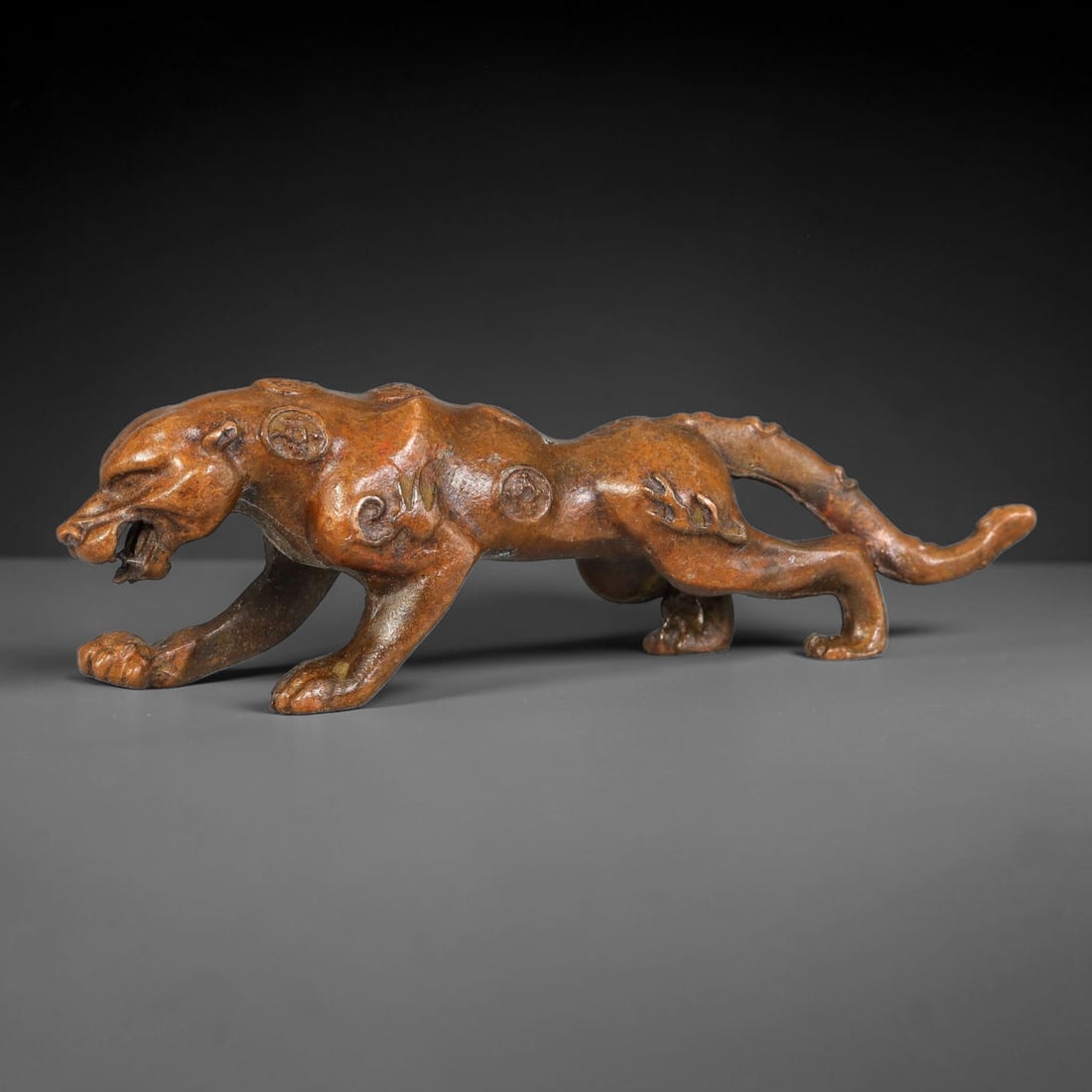 A Chinese Bronze Archaic Style Prowling Panther Effigy Figurine, 32AX: Cast bronze panther effigy captured in a low prowling stance with mouth agape, elongated body and extended tail. The torso is decorated with raised circular and scroll motifs, finely moulded to follow