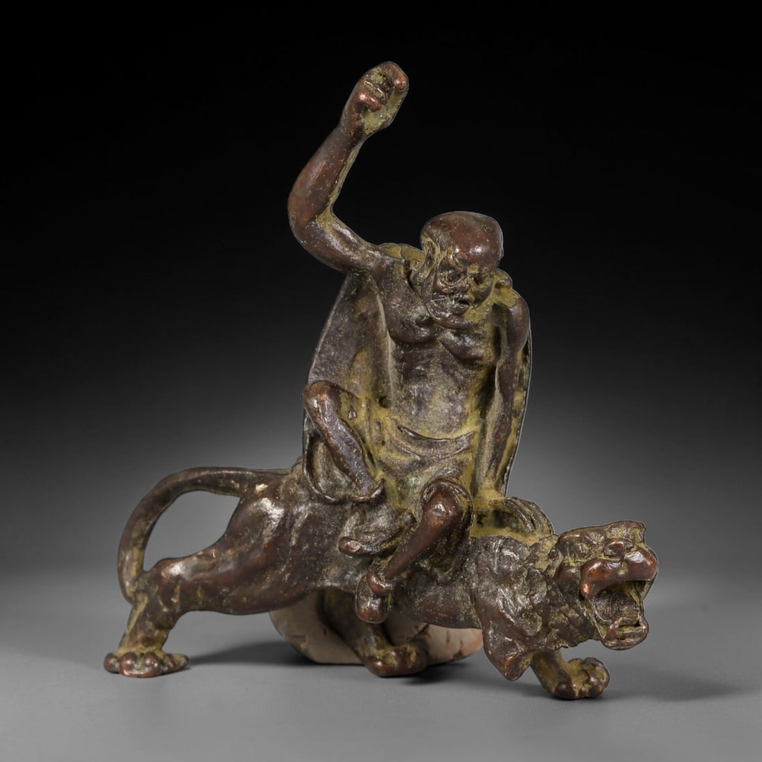 A Chinese Bronze Luohan Subduing Tiger Figural Group, 39AX: Cast bronze figural group depicting a luohan seated atop a roaring tiger, the arhat with one arm raised in a dynamic gesture, finely rendered facial features and textured robe detailing; the tiger mod