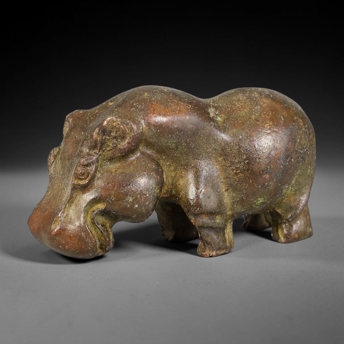 A Chinese Bronze Archaic Style Hippopotamus Zoomorphic Figurine, 38AX: Cast bronze zoomorphic figure modelled as a hippopotamus in a low, compact stance with rounded torso, broad muzzle and stylized facial detailing. The surface retains a heavy, multi-tonal aged patina w
