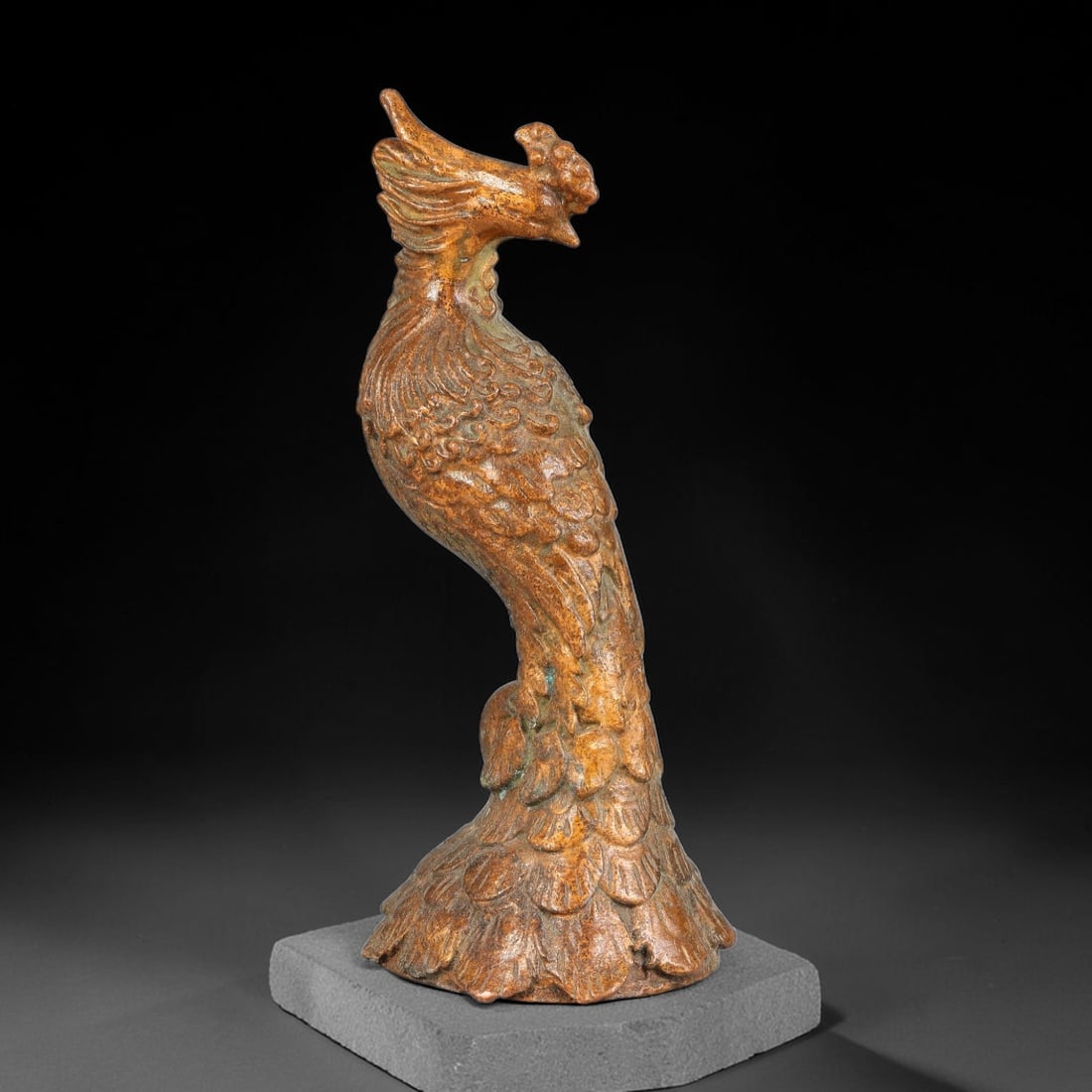 A Chinese Bronze Phoenix Effigy, 60AX: Cast bronze effigy modelled as a standing phoenix with crested head and gracefully curved neck. The body and base are rendered with layered feather relief detailing. The bronze surface carries warm ag