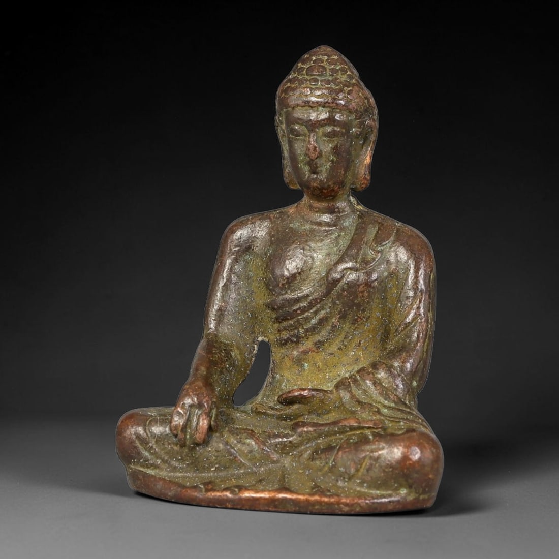 A Chinese Bronze Seated Buddha Figurine, 29AX: Cast bronze Buddha seated in lotus position, rendered with a rounded ushnisha, closed serene eyes and robe folds draped over one shoulder. The bronze bears a mottled aged patina with brown and green s