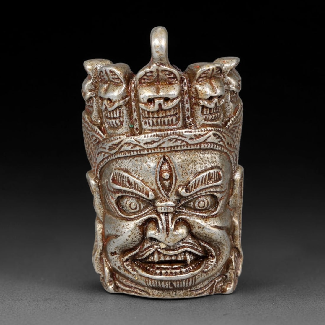 A Himalayan Tibetan Silvered Mahakala Head Pendant, 70BD: Cast silver pendant modelled as a wrathful Mahakala head crowned with a garland of skulls. The reverse features engraved Tibetan mantra script and a suspension loop at the apex. The silver exhibits an