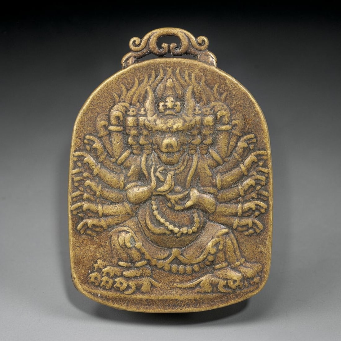 A Himalayan Bronze Yamantaka Relief Amulet Pendant, 69BD: Cast bronze amulet pendant with ornate scrolling suspension loop at top, featuring relief carving of multi-armed Yamantaka surrounded by flame mandorla. The piece bears warm aged bronze patina and fin