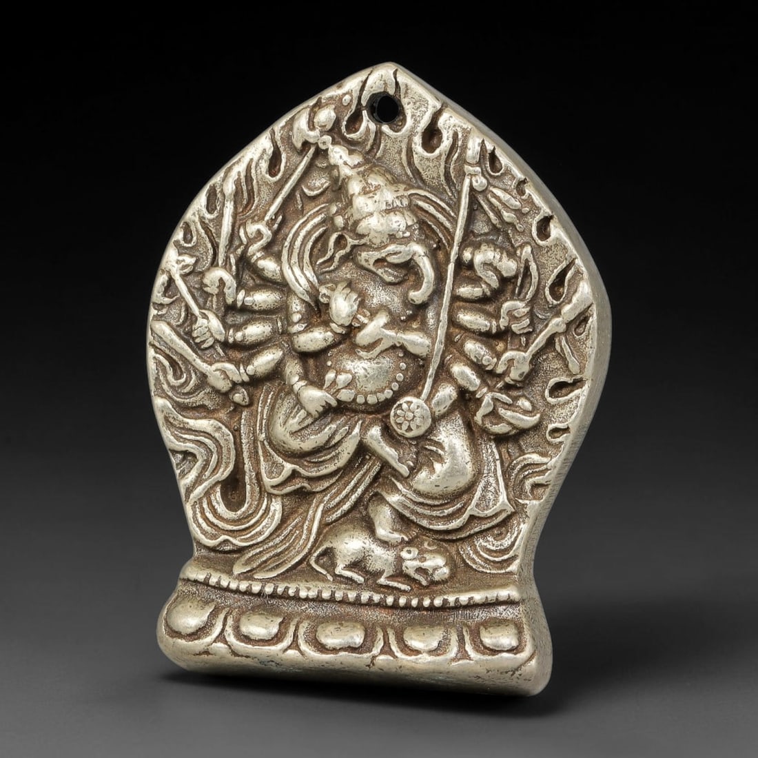 A Tibetan Silvered Bronze Ganesha Mandala Amulet Pendant, 68BD: Silvered bronze amulet pendant depicting a multi-armed Ganesha in dynamic relief, set within a flame-arched mandala frame with a pierced suspension hole at the top. The reverse is engraved with a stup