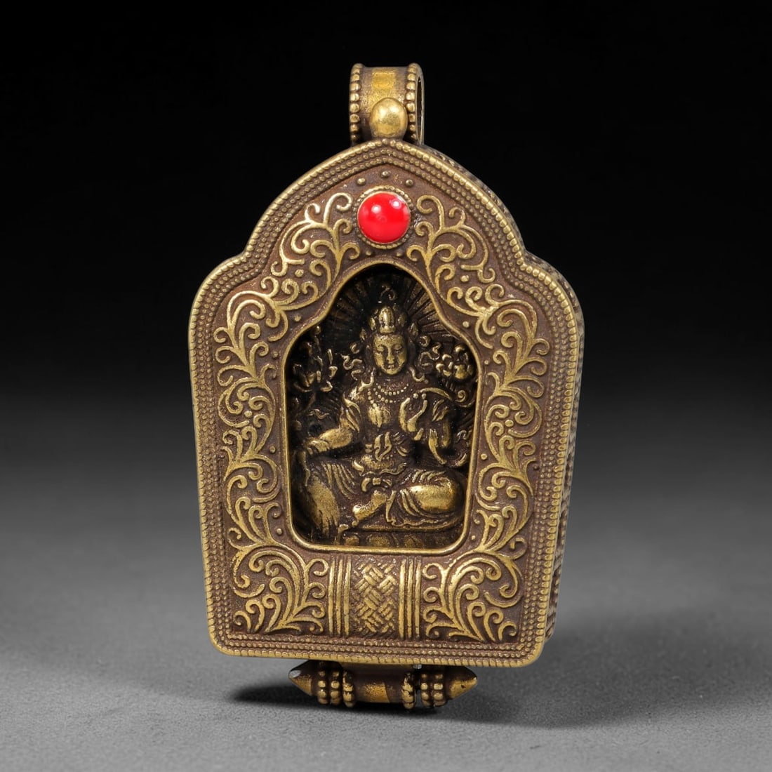 A Himalayan Brass White Tara Ghau Shrine Pendant, 76BD: Cast brass portable ghau shrine pendant with arched top and decorative scrollwork borders, set with a single red cabochon at the apex. The central niche holds a relief carving of White Tara seated in 