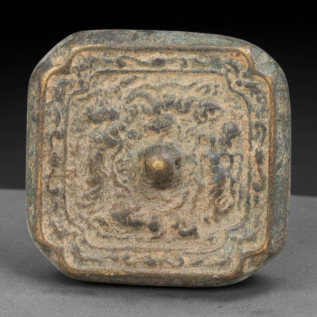 A Chinese Square Bronze Mirror, 62BD: Small cast bronze square mirror with rounded corners, featuring a central hemispherical knob. The face is decorated with relief scrolling mythical creature motifs enclosed within a double border frame