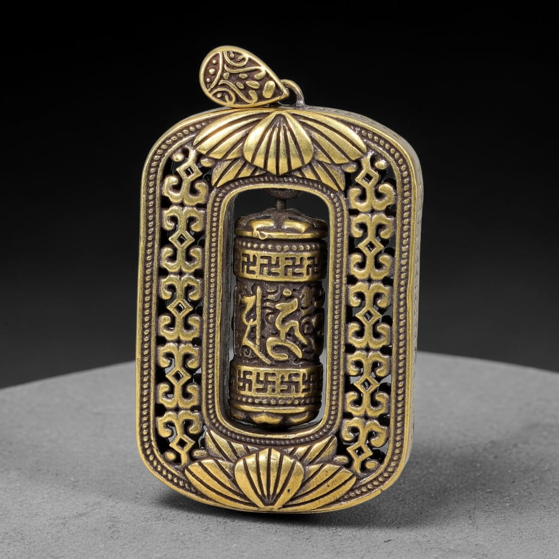 A Himalayan Brass Openwork Prayer Wheel Ghau Pendant, 55BD: Cast brass ghau pendant with an openwork rectangular frame decorated with scrolling auspicious motifs and lotus carvings at top and bottom. A freely rotating miniature prayer wheel engraved with Tibet