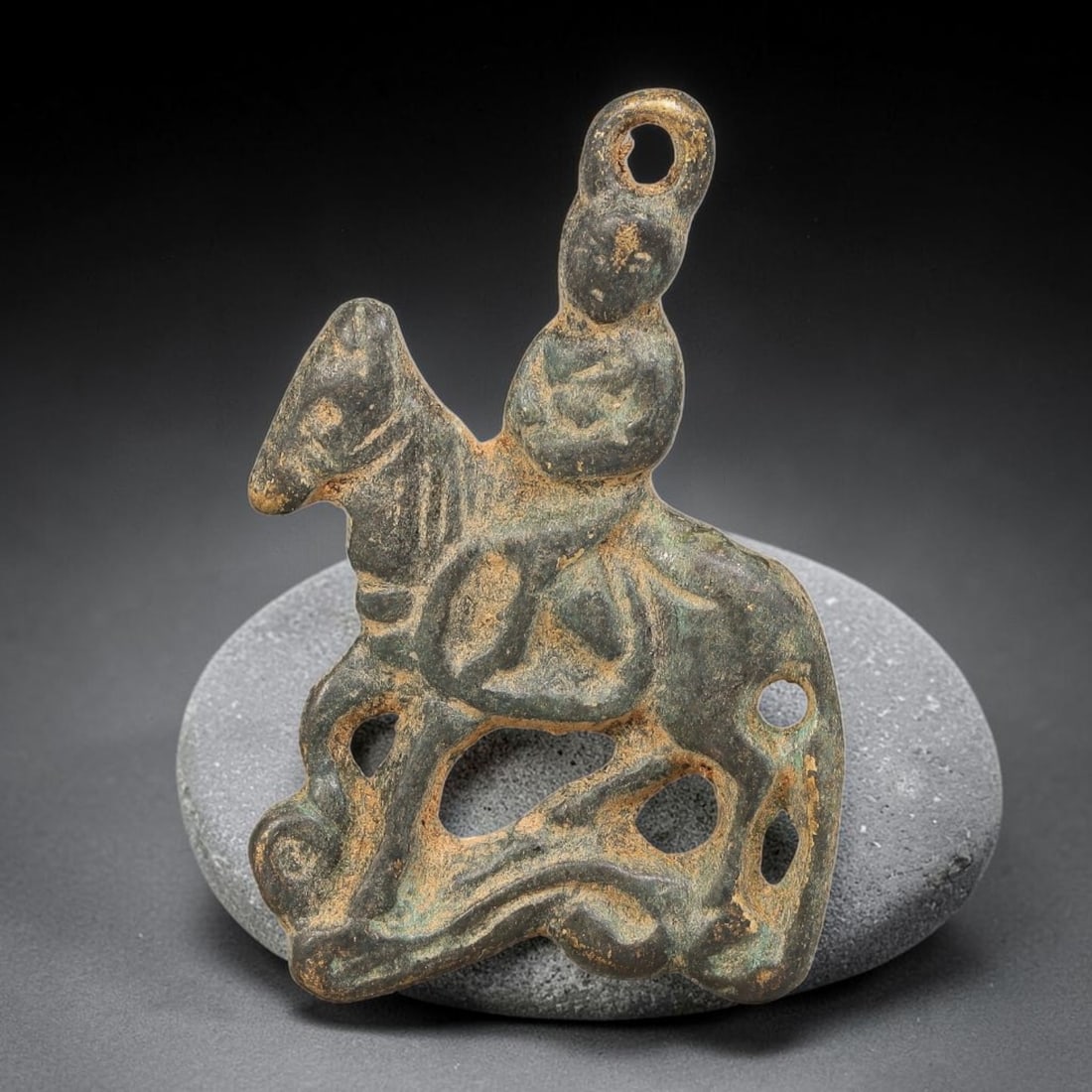 A Chinese Bronze Openwork Rider on Horse Amulet Pendant, 60BD: Cast bronze openwork amulet pendant depicting a seated rider upon a horse, with a suspension loop formed at the apex. The piece features pierced cutout detailing across the base, with heavy burial pat