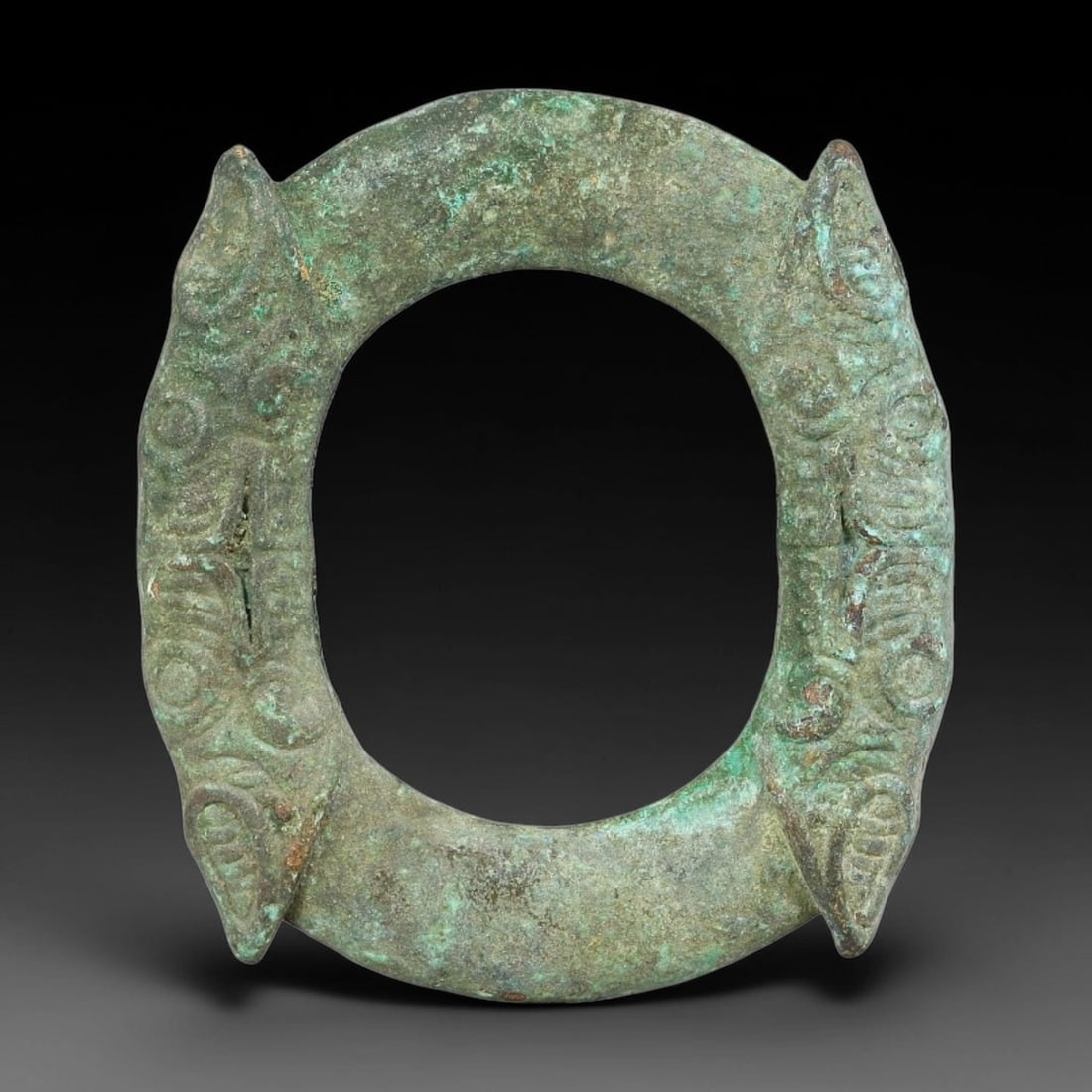 A Chinese Bronze Twin Beast Head Amulet Ring, 57BD: Cast bronze amulet ring decorated with twin beast head relief motifs positioned on opposite sides of the hoop. The surface displays thick green burial patina and textured corrosion across the raised c