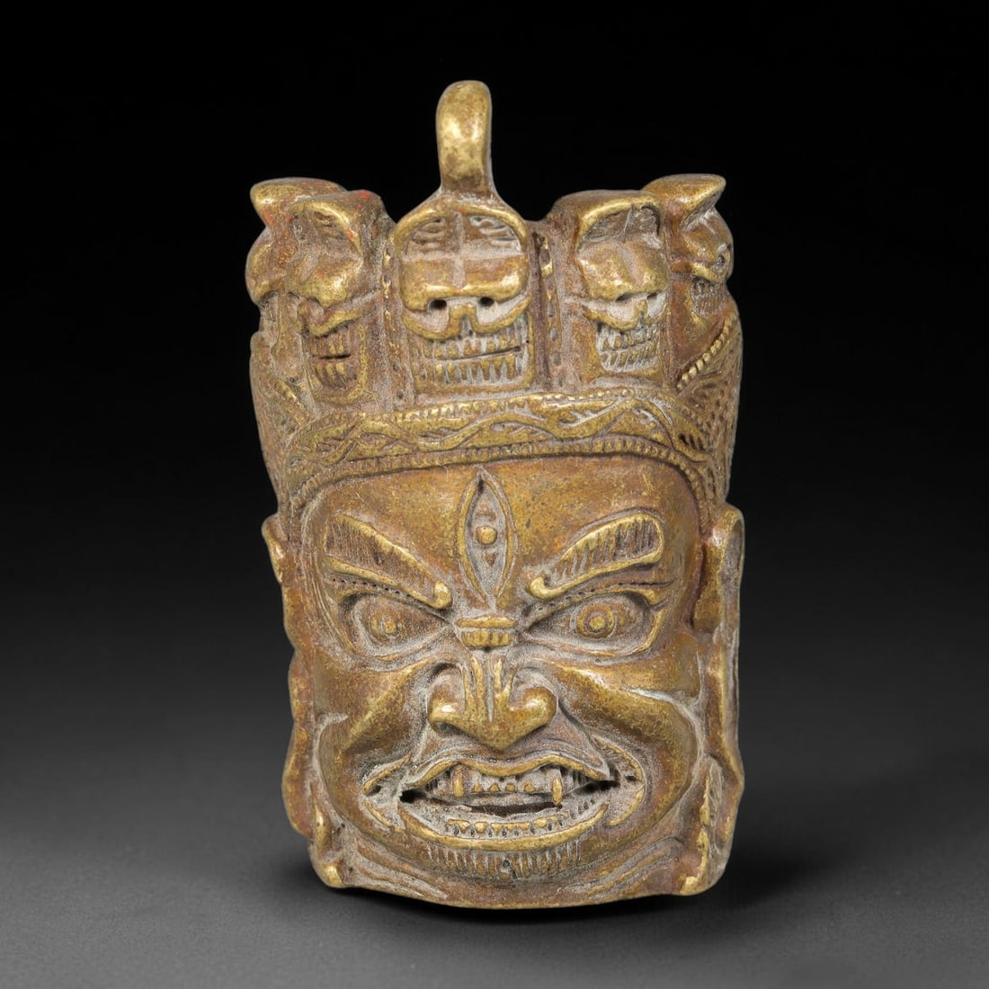 A Himalayan Tibetan Bronze Mahakala Head Pendant, 71BD: Cast bronze pendant modelled as a wrathful Mahakala head crowned with skull motifs. The reverse bears engraved Tibetan mantra script and a suspension loop at the apex. The bronze has a warm aged golde
