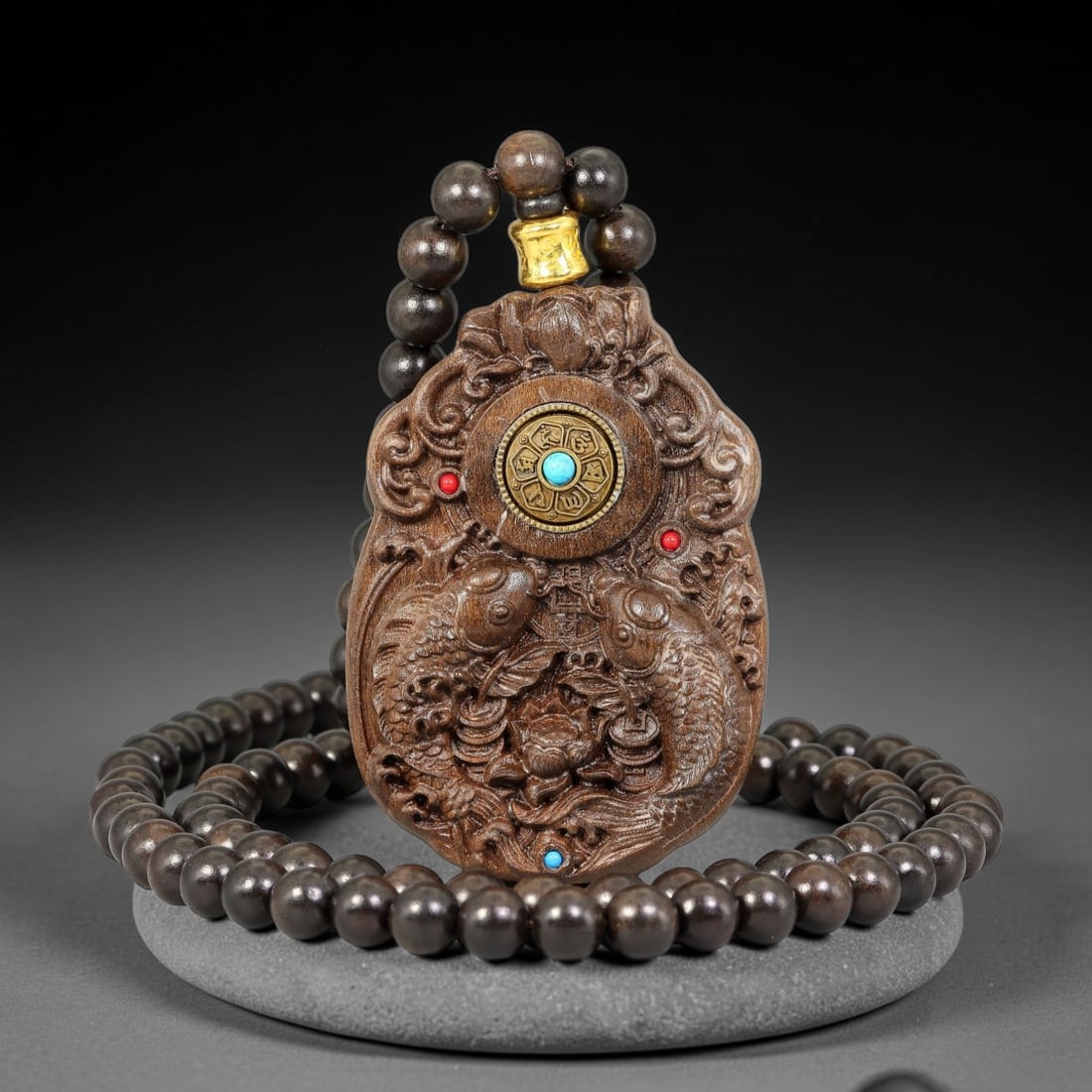 A Chinese Carved Wood Twin Carp Rotating Dharma Wheel Pendant with Beaded Necklace, 86BD: Plaque-form carved wood pendant with scrolling lotus and cloud ornamentation at the upper register, deeply relief carved with paired carp amid lotus and water motifs. A rotatable metal dharma wheel se