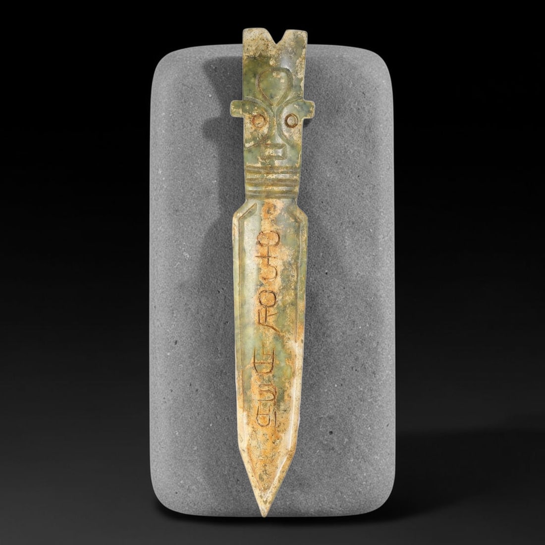 A Chinese Archaistic Green Jade Dagger with Mask Head Pommel, 88AX: Elongated dagger-form jade carving topped with a stylized archaic mask pommel with circular eyes and incised facial features. Horizontal banding decorates the neck, while the blade bears incised archa