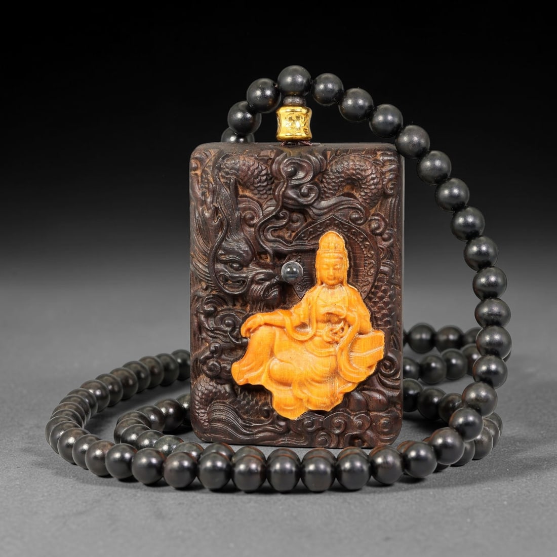 A Chinese Carved Wood Plaque Pendant with Guanyin Inlay and Beaded Necklace, 85BD: Rectangular carved wood plaque pendant deeply relief carved with swirling dragon and cloud motifs, featuring an inlaid seated Guanyin figure at the centre and a small inset cabochon detail. Supplied w