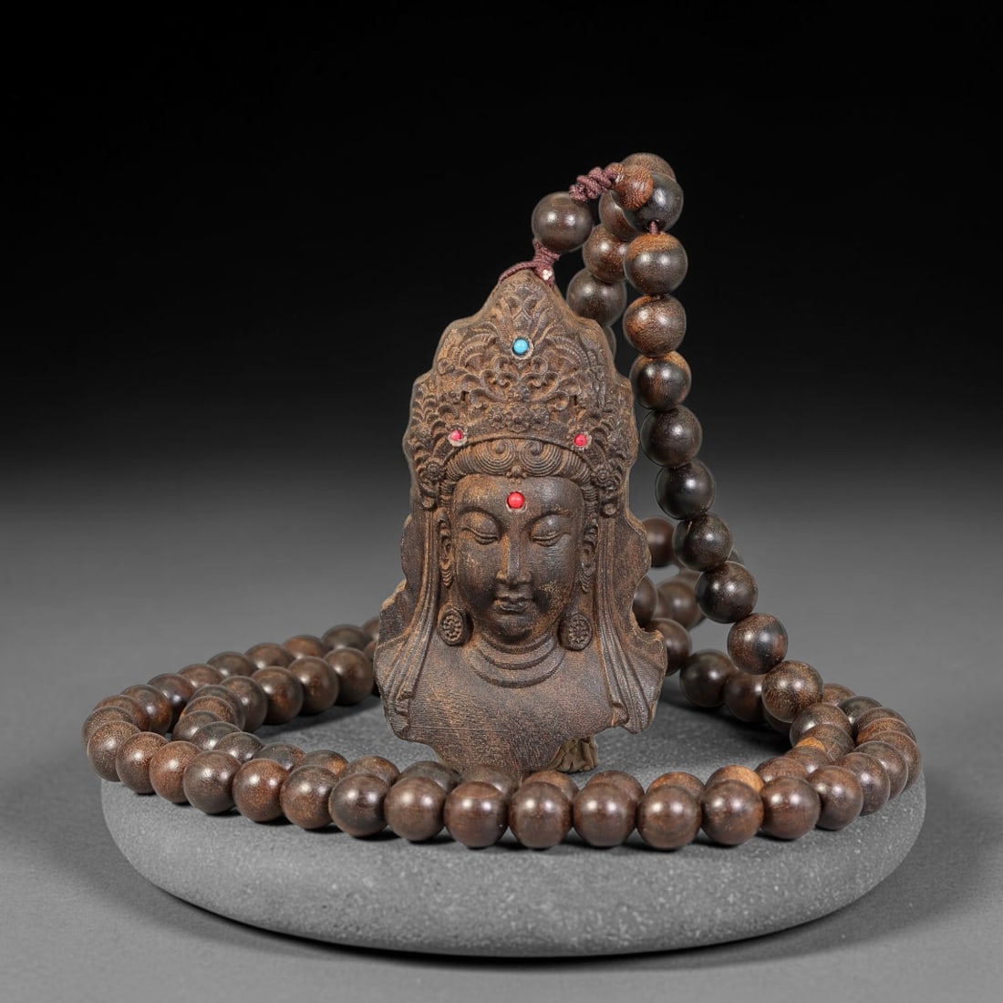 A Chinese Carved Wood Guanyin Head Pendant with Beaded Necklace, 81BD: Carved wood pendant finely modelled as a crowned Guanyin head, adorned with inset coloured stone accents upon the headdress and forehead. The pendant is strung with a full strand of matching polished 