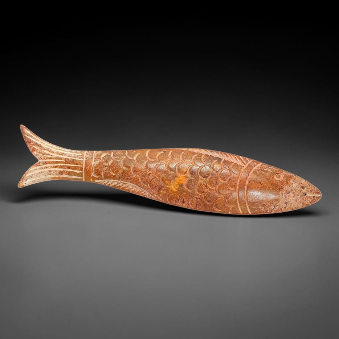 A Chinese Carved Reddish-Brown Jade Fish Pendant, 95AX: Elongated jade piece fully sculpted in the form of a fish, with fine incised lines rendering overlapping scales, dorsal, pectoral and tail fins. A small suspension hole is drilled at the tip of the mo