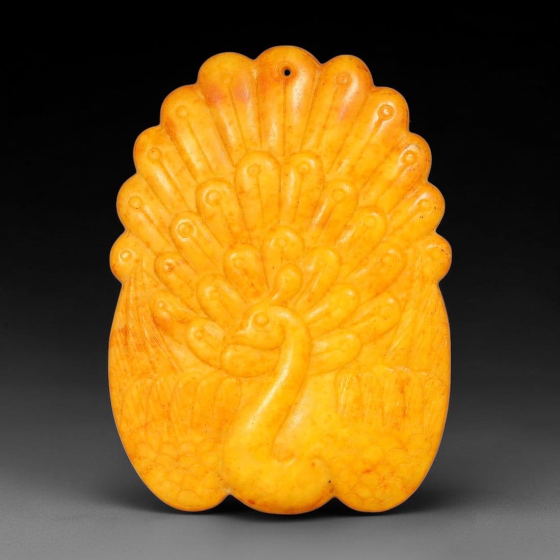 A Chinese Carved Yellow Jade Peacock Plaque Pendant, 97AX: Solid relief-carved jade plaque depicting a peacock with fully fanned tail feathers, each feather detailed with incised ocellus motifs. A small suspension hole is drilled at the upper edge of the plaq