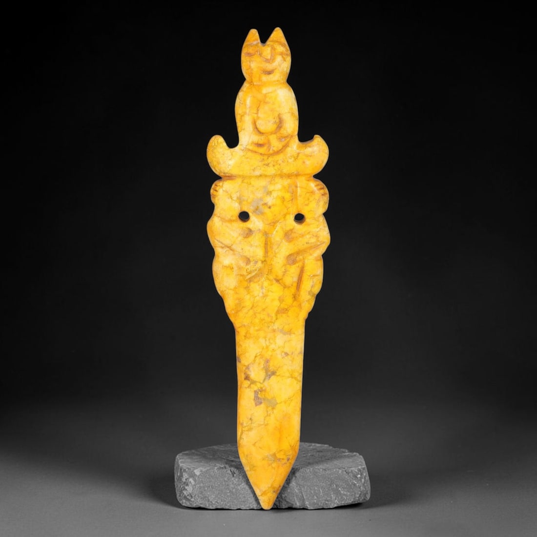 A Chinese Carved Yellow Jade Dagger Pendant with Anthropomorphic Finial, 93AX: Elongated dagger-form jade pendant topped by a stylized seated anthropomorphic figure. The midsection bears relief carved beast mask ornamentation with two drilled piercing holes. The blade tapers smo