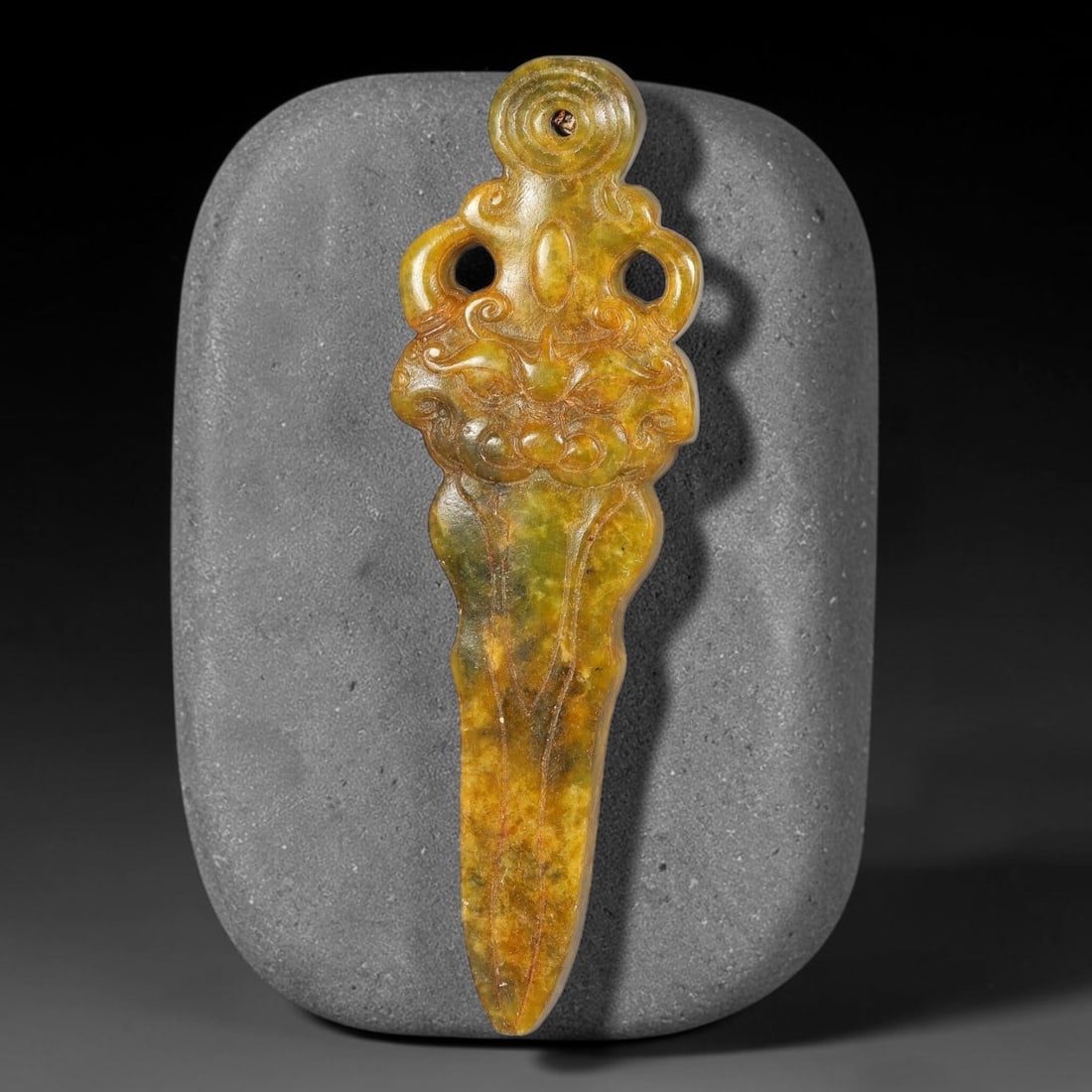 A Chinese Carved Mottled Jade Dagger Pendant, 36BD: Dagger-form jade pendant topped with a circular suspension ring. The upper section is relief carved with an ornate beast mask flanked by curling scroll motifs and pierced openwork cut-outs. The tapere