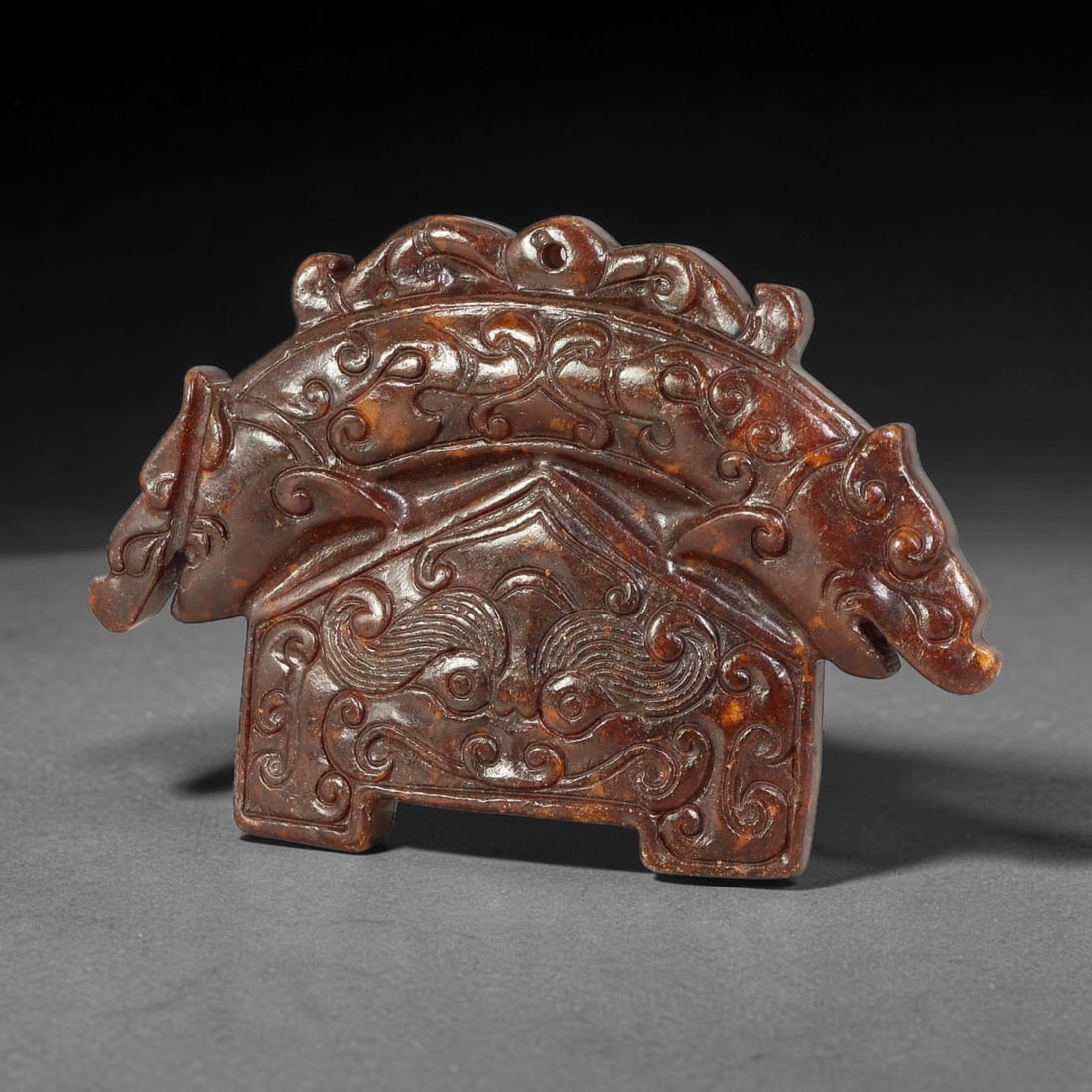 A Chinese Carved Dark Jade Twin Dragon Arch Pendant, 19BD: Arch-form dark jade pendant with dragon heads projecting outward at each terminal, the upper curved surface decorated with continuous scroll relief carving. The lower rectangular section bears a centr