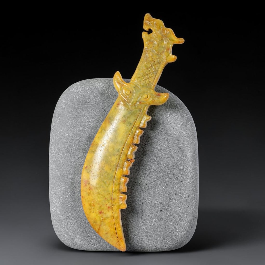 A Chinese Carved Mottled Yellow Jade Curved Blade Pendant with Dragon Head Pommel, 89AX: Curved saber-form jade pendant crowned by a stylized dragon head pommel, with incised lattice detailing along the handle. The guard is formed by a symmetrical beast mask motif, and the outer blade edg