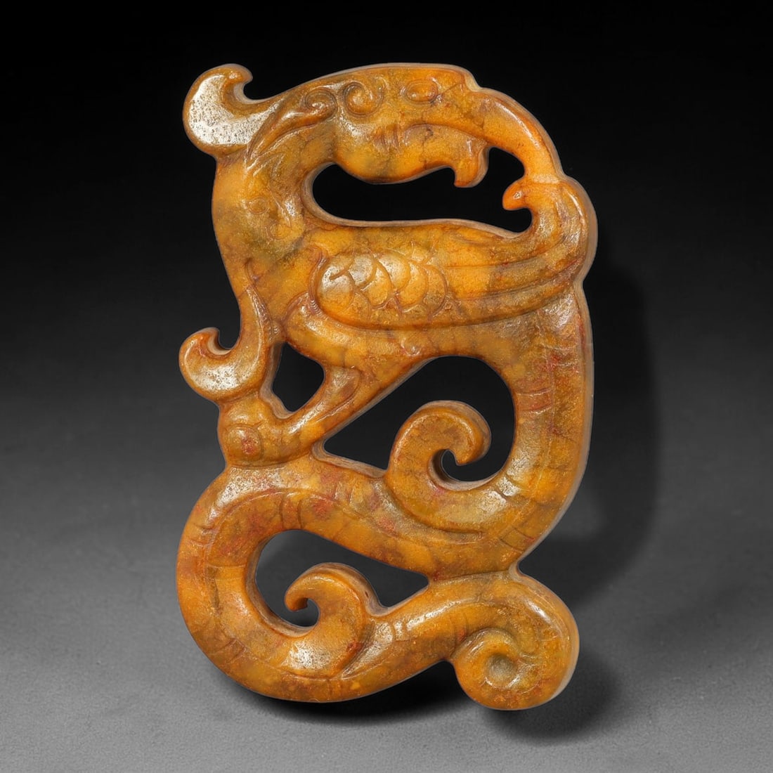A Chinese Openwork Yellow Jade Coiled Phoenix Pendant, 31BD: Openwork yellow jade pendant carved as a dynamically coiled phoenix, with stylized feathering, curled crest and swirling tail elements defined by incised lines. Fluid pierced negative space winds thro
