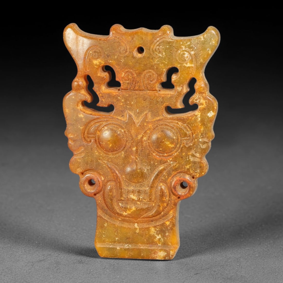 A Chinese Openwork Carved Jade Anthropomorphic Mask Pendant, 16BD: Openwork carved jade mask pendant modelled with prominent circular eyes, ornate headdress and scroll ornamentation. Pierced suspension hole at the apex, with additional cut-out details flanking the fa