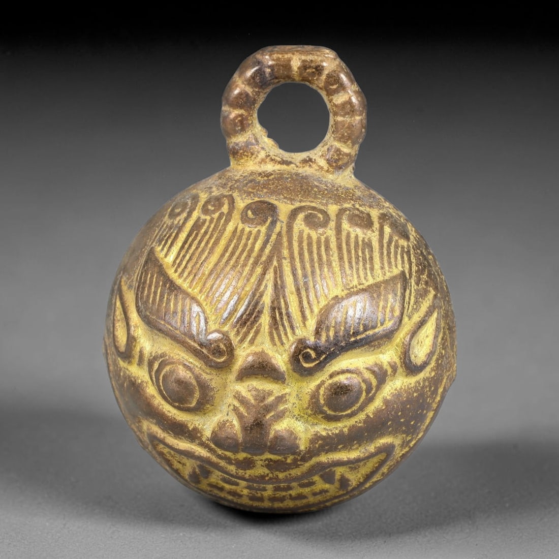 A Chinese Bronze Guardian Beast Bell, 51BD: Cast bronze spherical bell decorated in relief with a detailed guardian beast face, featuring defined eyes, brows and open mouth with visible teeth. A textured suspension loop rises from the apex for 
