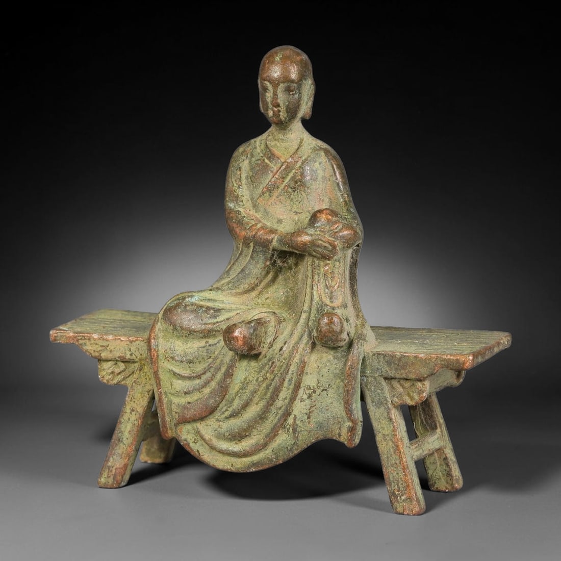 A Chinese Bronze Bench Seated Luohan Sculpture, 53AX: Cast bronze sculpture depicting a luohan seated upon a rustic bench. The figure wears flowing layered monastic robes with naturalistic drapery folds, holding a rounded object in the hands. The surface