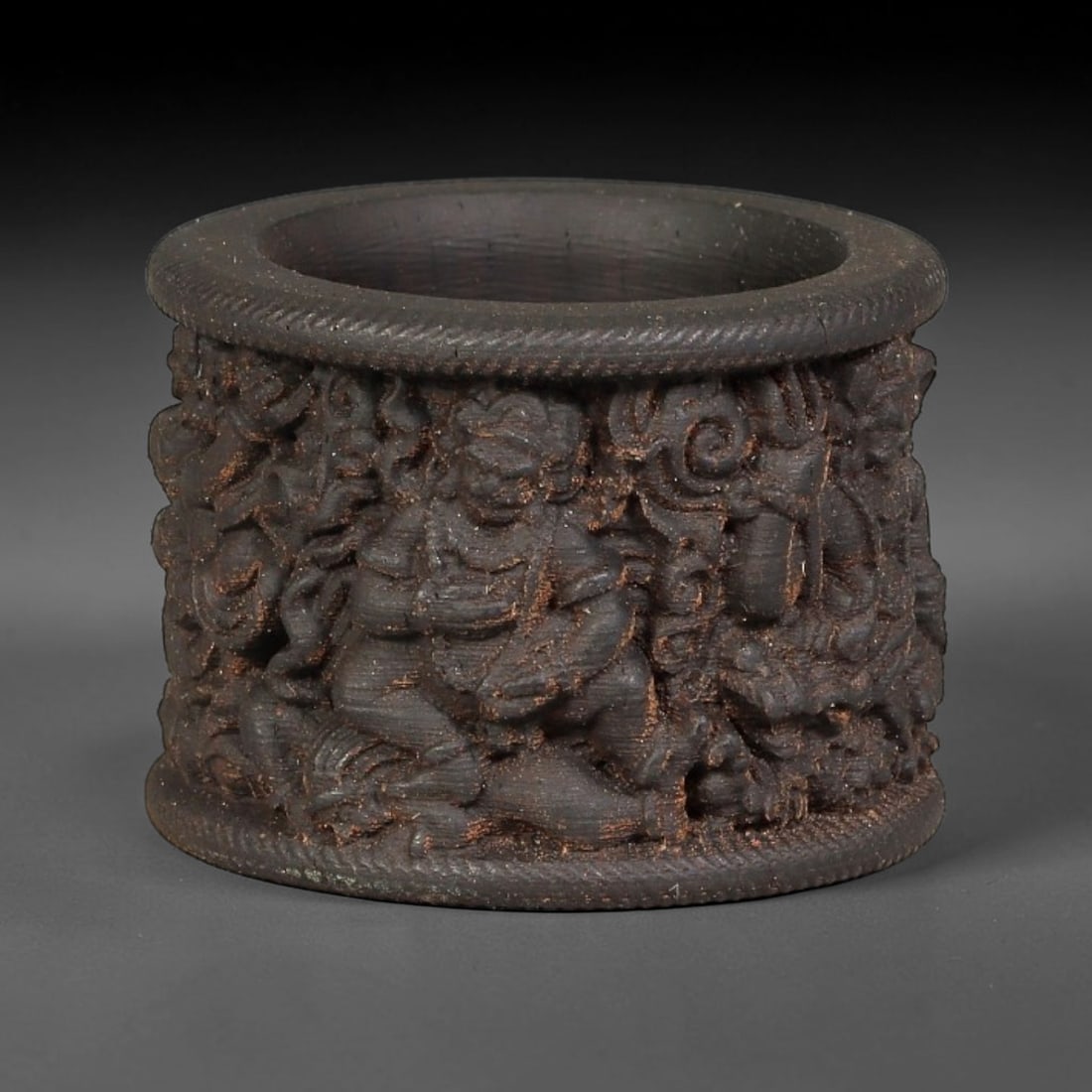 A Chinese Agarwood Carved Thumb Ring, 2BF: Deeply relief-carved agarwood thumb ring, densely decorated with figural and scrolling motifs encircling the exterior. The top and bottom rims are finished with twisted rope-style border carving. Fine