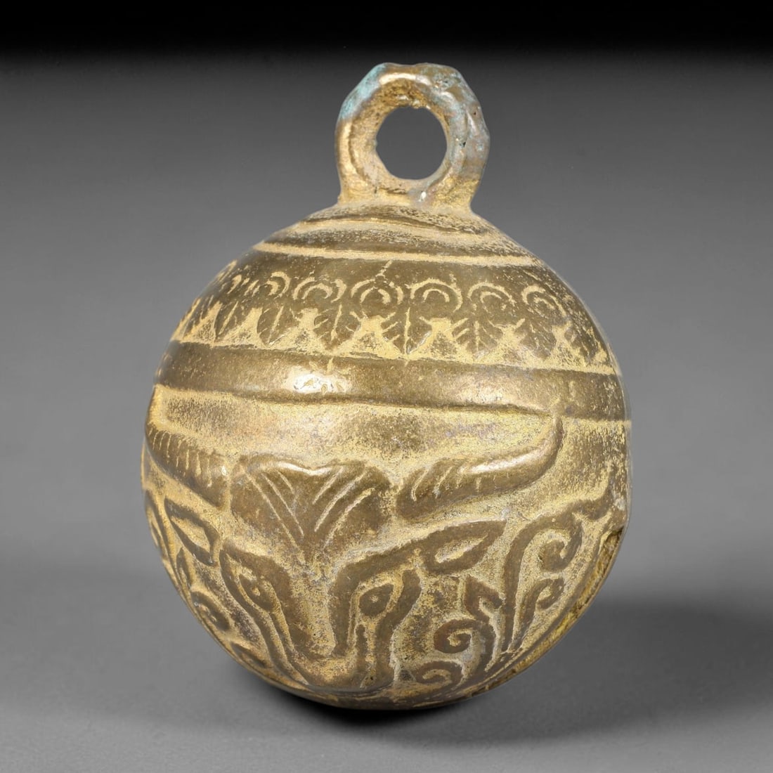 A Chinese Bronze Ox Head Bell, 52BD: Cast bronze spherical bell decorated in relief with an ox head motif, paired with horizontal bands and scrolling foliate ornament encircling the upper body. A heavy suspension loop is formed at the to