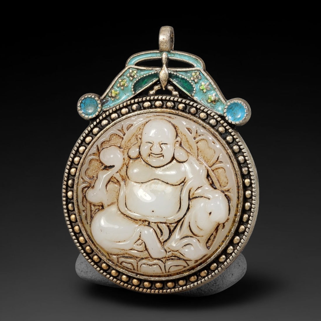 A Chinese Enameled Metal Mounted Carved Budai Double-Sided Pendant, 3BF: Double-sided circular pendant with a carved pale stone plaque depicting seated Budai holding a ruyi sceptre on one face, set within a beaded metal frame. The reverse features raised cloisonne-style en