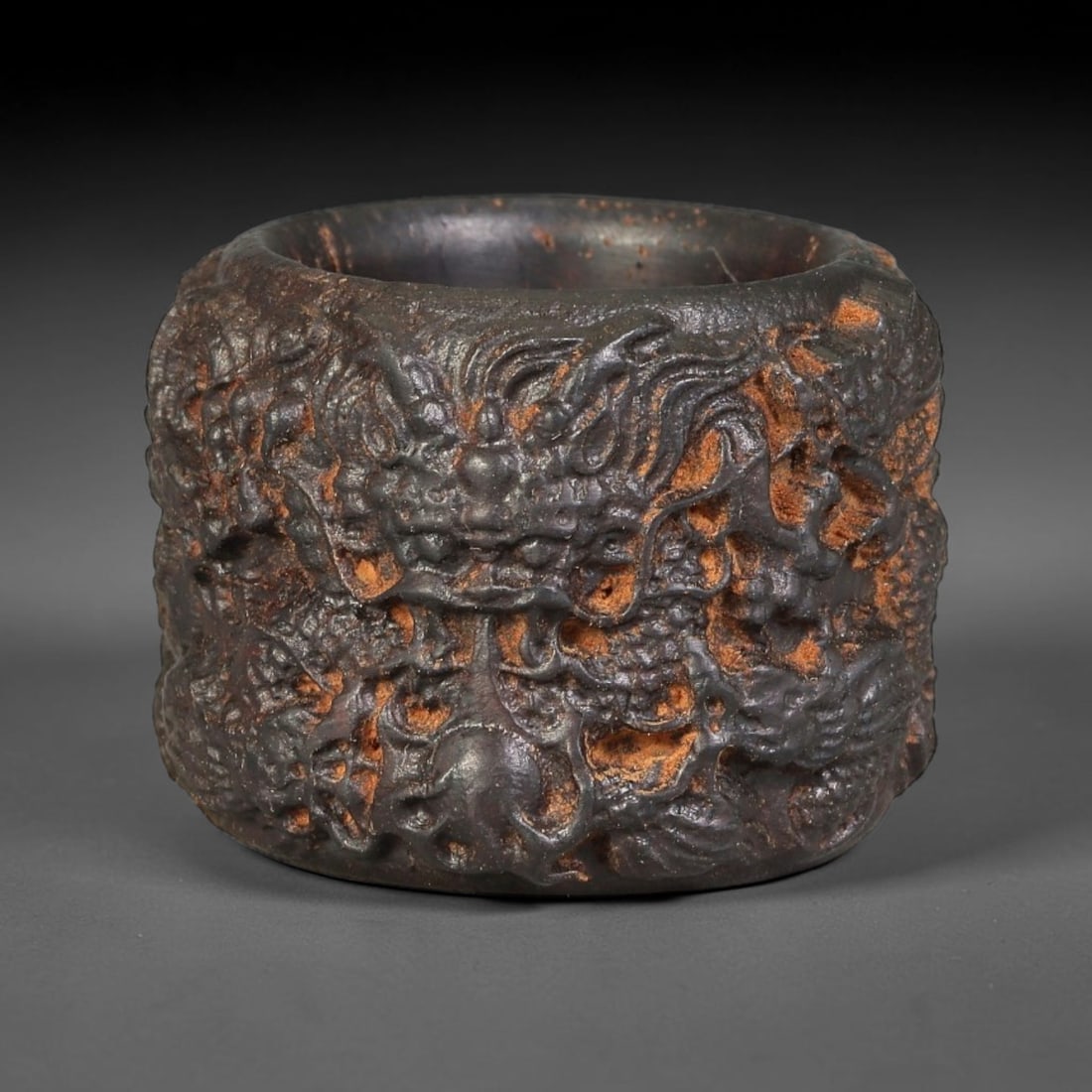 A Chinese Agarwood Carved Dragon Thumb Ring, 100BD: Deep relief agarwood thumb ring fully encircled by a dynamic dragon motif chasing a flaming pearl. The carving features deeply undercut details, revealing patches of warm reddish-brown wood against th