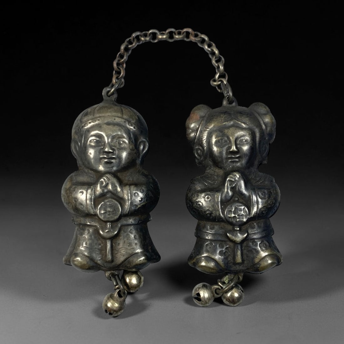 A Chinese Vintage Silver Boy & Girl Figural Pendant Set, 96BD: Vintage silver pendant set depicting a paired boy and girl traditional child figures, connected by a delicate link chain. Each figure stands with hands clasped, adorned with embossed costume details a
