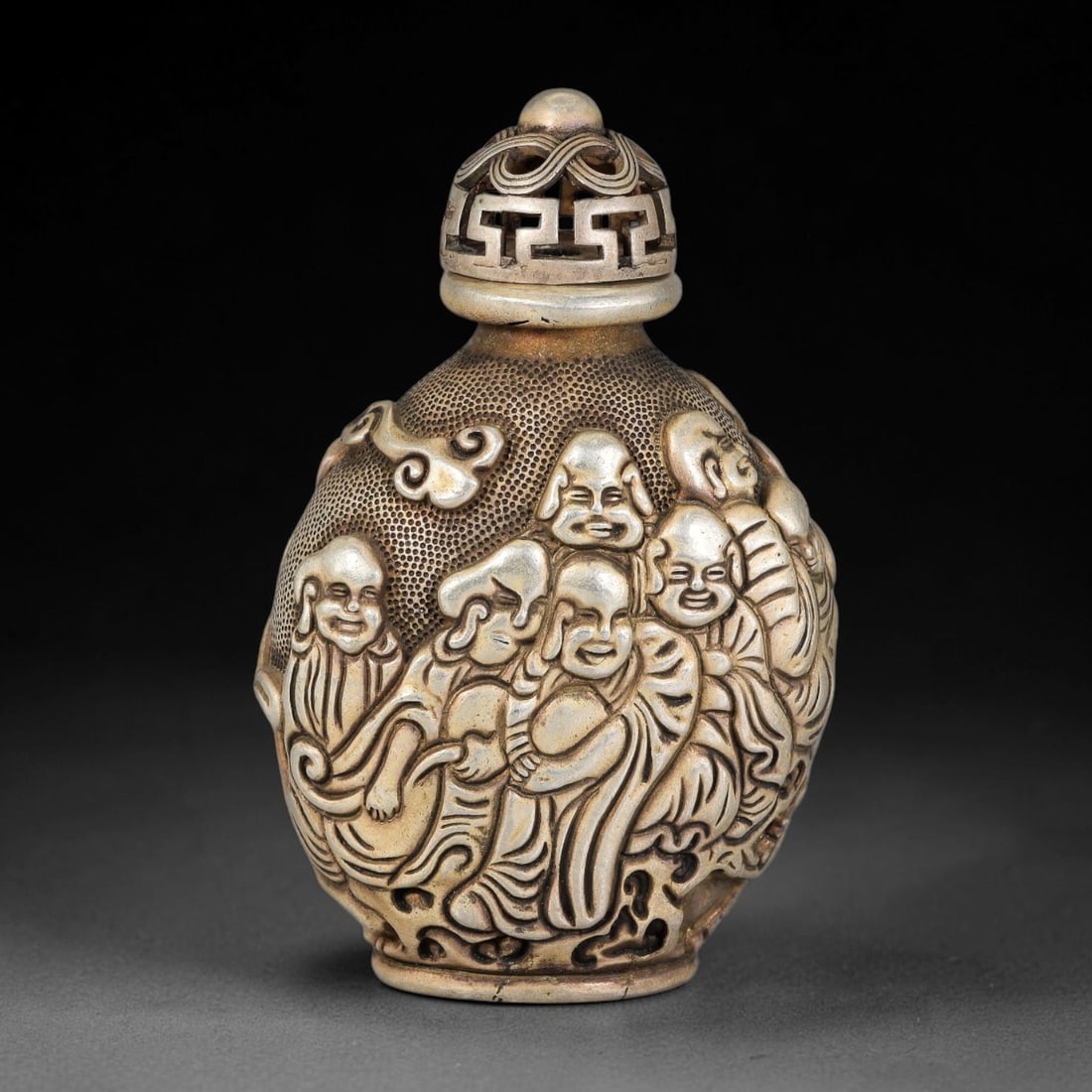 A Chinese Relief Metal Snuff Bottle with Eighteen Luohan Figures, 21AV: Ovoid form metal snuff bottle heavily decorated in high relief depicting multiple luohan set against a stippled textured ground. It is fitted with an openwork pierced stopper with geometric fretwork p