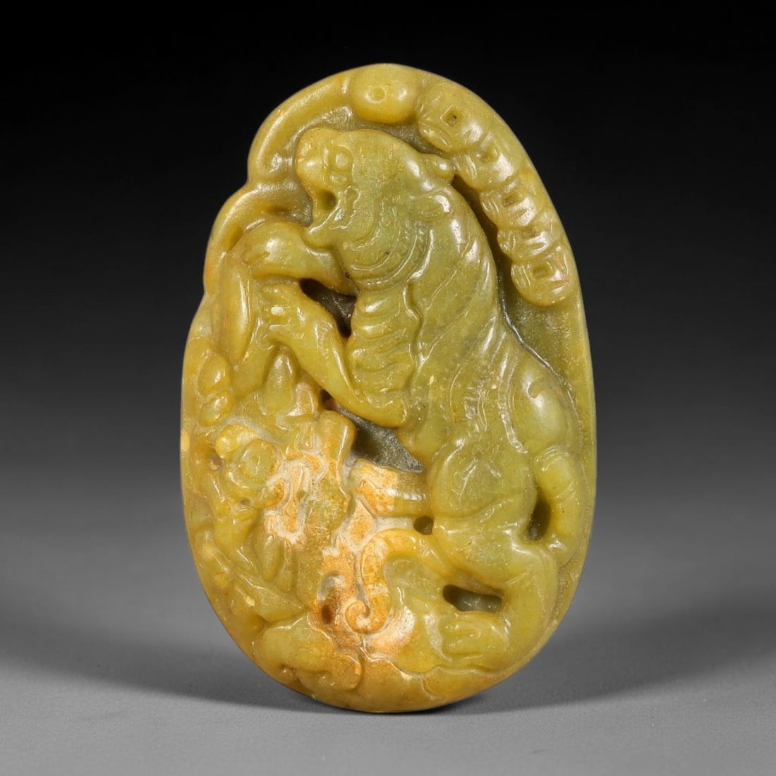 A Chinese Carved Jade Tiger Plaque Pendant, 27BD: Oval jade plaque with relief and openwork carving depicting a fierce tiger amid rocky and cloud motifs. The stone exhibits an olive-yellow tone with areas of lighter weathered patina. The upper edge i