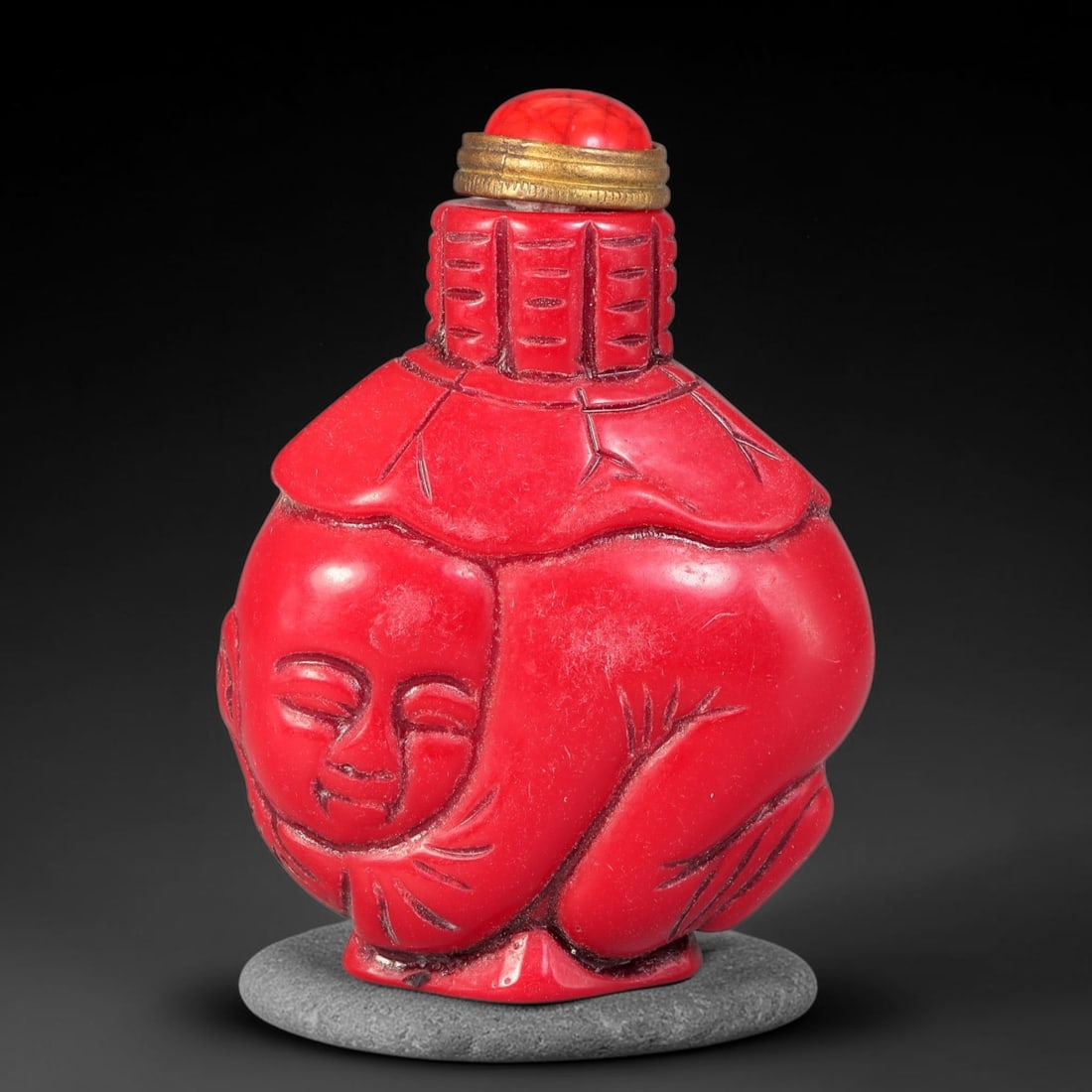 A Chinese Red Resin Carved Child Figural Snuff Bottle, 7AX: Figural snuff bottle crafted from red resin, carved in the form of a curled child with serene closed-eye features, draped fabric and textured detailing on the neck. It is fitted with a red domed stopp