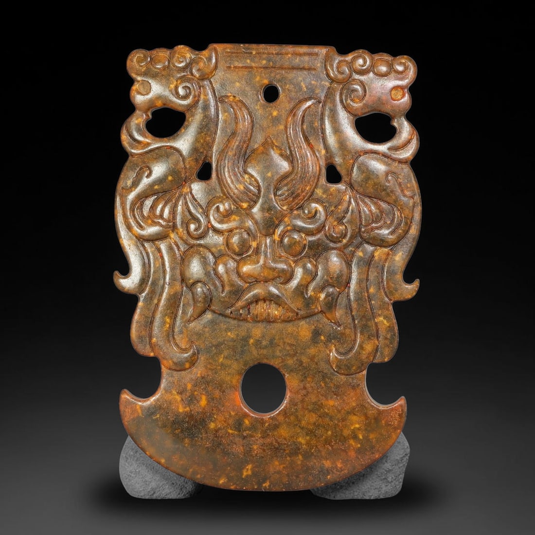 A Chinese Openwork Mottled Dark Jade Axe Plaque with Beast Mask and Twin Dragons, 100AX: Axe-shaped openwork jade plaque centered on a prominent relief-carved archaic beast mask, flanked by symmetrical stylised dragon figures with curling scroll forms and pierced voids. Two circular perfo