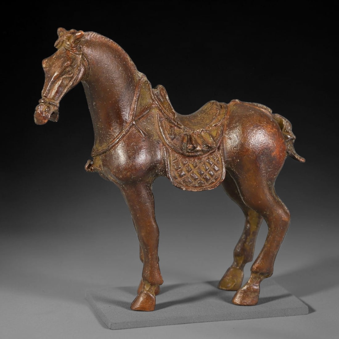 A Chinese Bronze Saddled Horse Figurine, 65AX: Cast bronze standing horse equipped with detailed saddle and bridle trappings, with defined musculature, neatly combed mane and stylized hooves. The bronze exhibits a rich warm brown aged patina, with