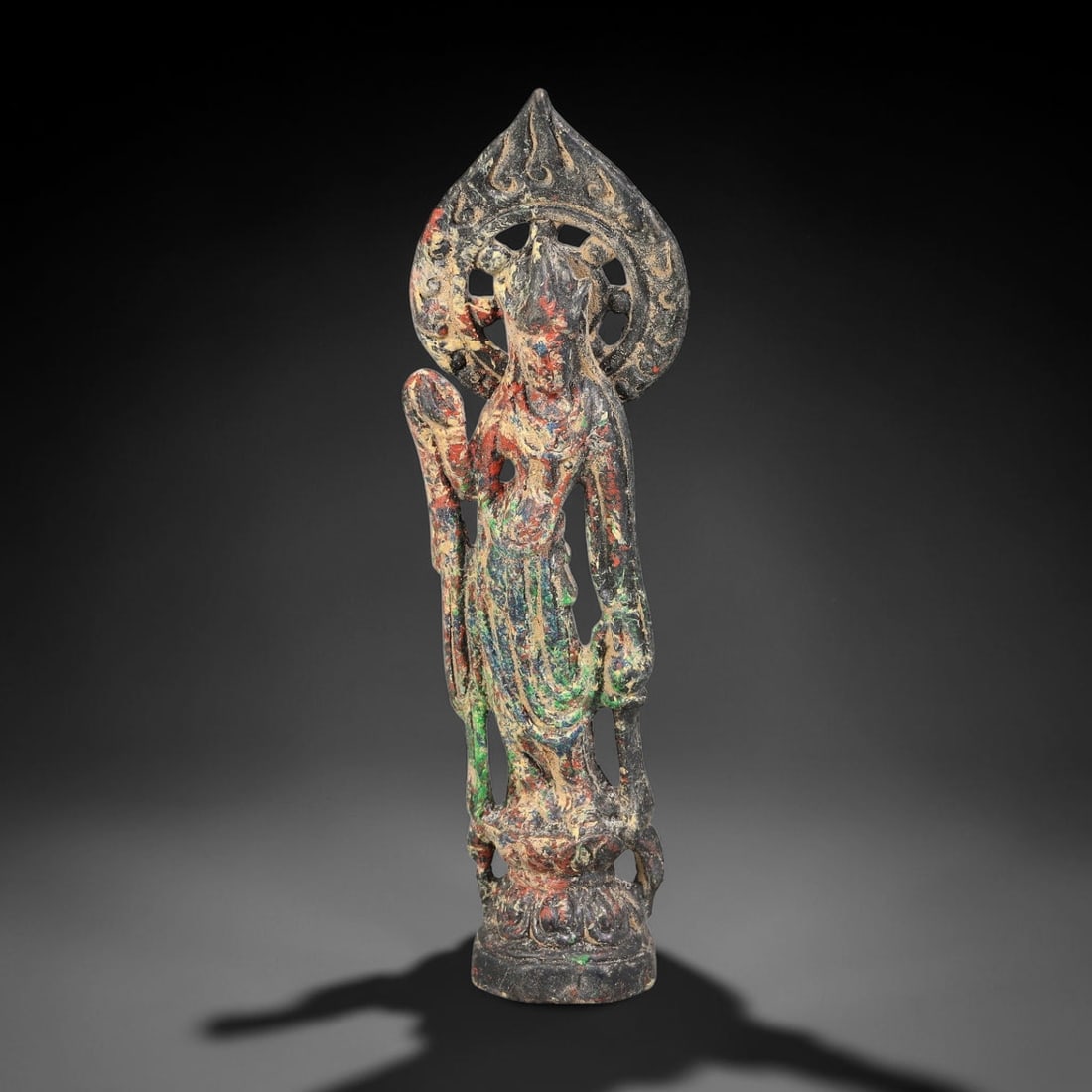 A Chinese Bronze Standing Guanyin with Painted Traces, 54BD: Cast bronze figure of standing Guanyin atop a small lotus pedestal, backed by a pierced flame mandorla. Remnants of polychrome pigment in red, green and blue tones remain across the robe and mandorla 