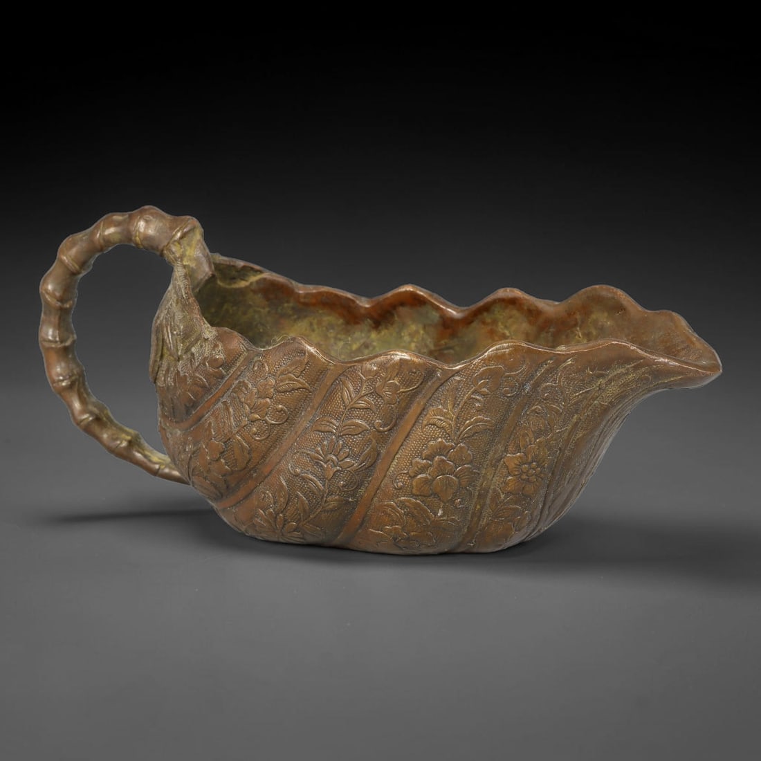 A Chinese Bronze Leaf-Form Pouring Vessel, 79AX: Cast bronze vessel modelled after a folded leaf with a wavy scalloped rim, fitted with a segmented bamboo-style handle. The exterior surface is densely decorated with raised relief floral sprays follo