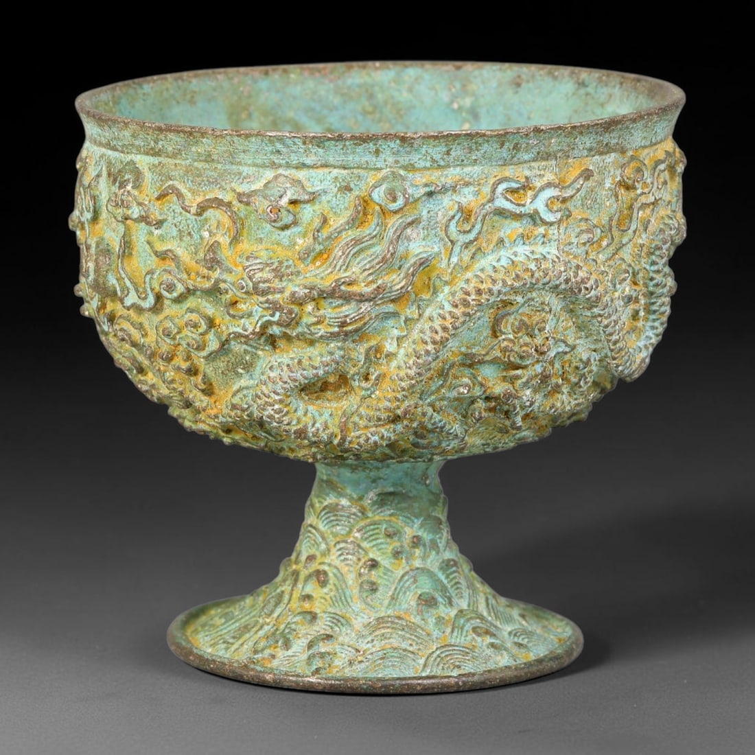 A Chinese Bronze Stem Cup with Dragon Relief Ornament, 99BD: Bronze stem cup decorated with raised relief coiled dragons and scrolling cloud patterns across the bowl. The tall stem is cast with stylized wave motifs. The surface is covered in mottled green and g