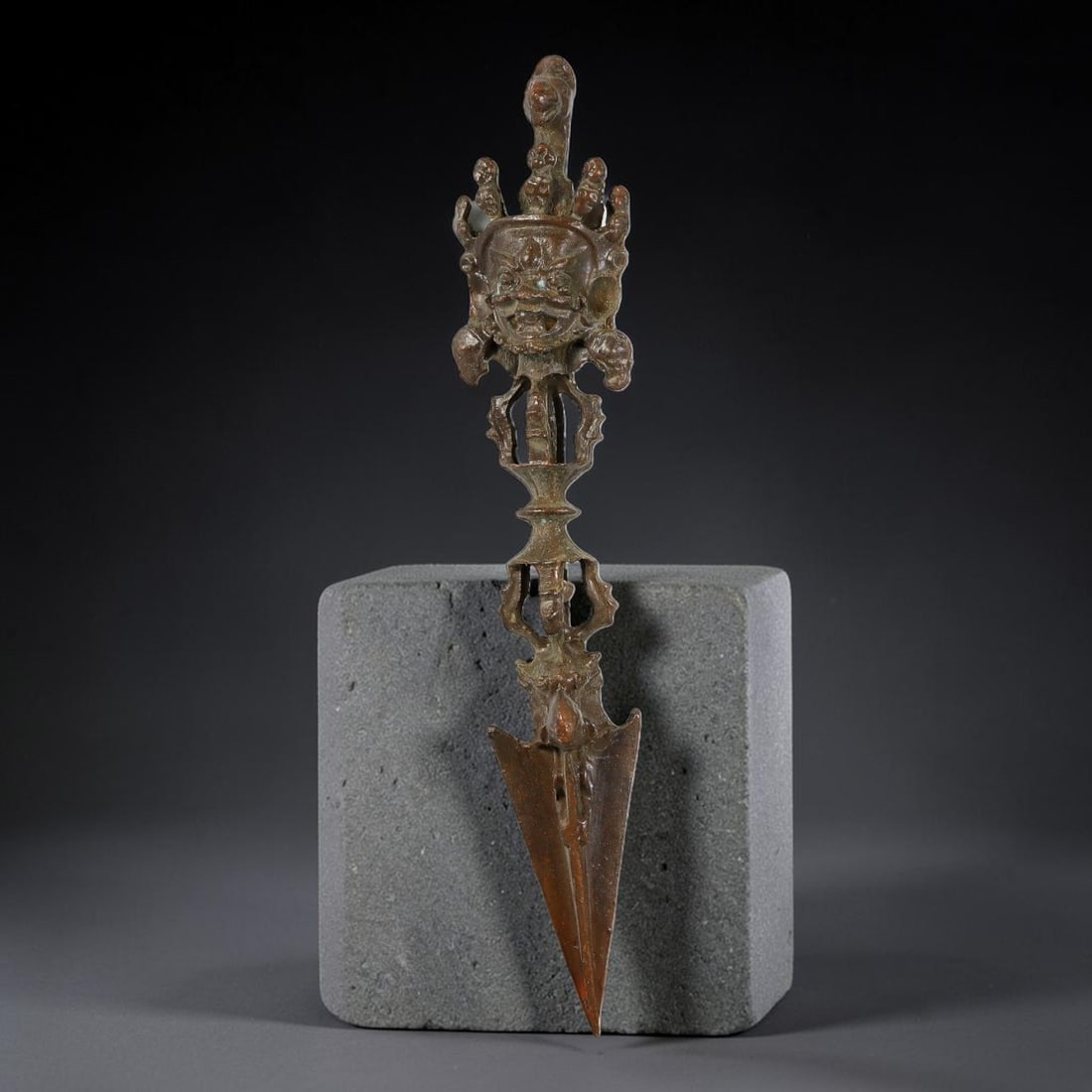 A Chinese Tibetan Openwork Bronze Phurba Ritual Dagger, 37AD: Cast in bronze, this ceremonial phurba is topped with a prominently modelled wrathful deity mask encircled by attendant small decorative figures. The shaft incorporates extensive pierced open-work sec