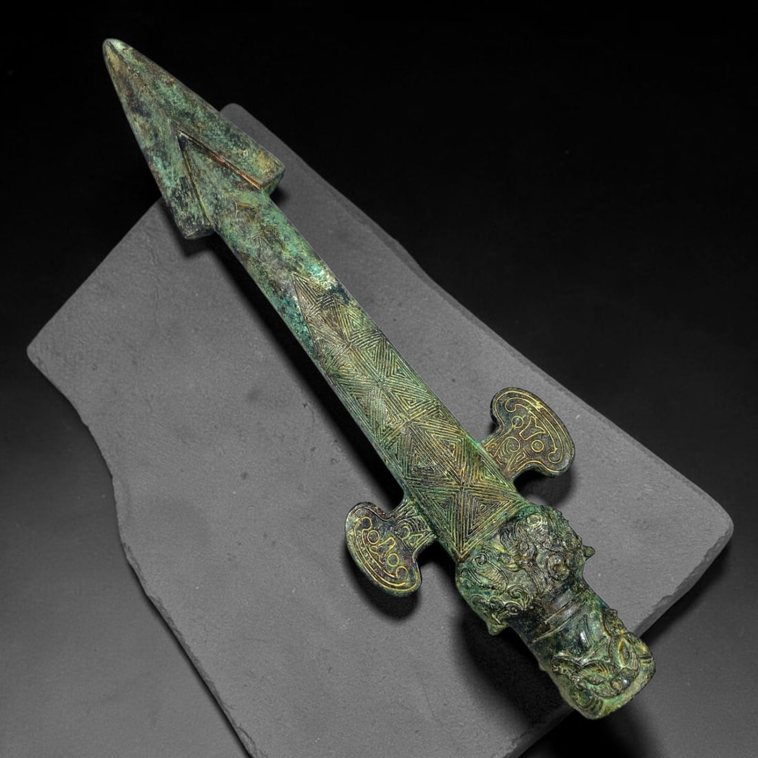 A Chinese Archaic Style Bronze Spear with Incised Geometric and Zoomorphic Decoration, 65BP: This archaic style bronze spear is finely decorated with dense incised geometric patterns running down the shaft, paired with stylized scroll shaped lugs and a detailed beast-head finial at the base. 