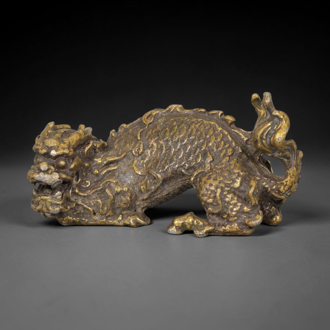 A Chinese Gilt Bronze Recumbent Dragon, 87BT: Cast in gilt bronze, the figure depicts a recumbent dragon with scaled body, stylized dorsal crests, defined facial features and a flowing curled tail. Remnants of gilding remain across the surface, w