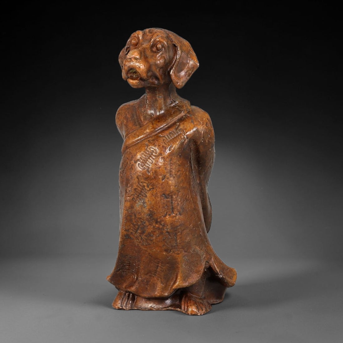 A Chinese Antique Bronze Anthropomorphic Zodiac Dog Figurine, 100BT: This bronze figurine portrays the zodiac dog with a dog head attached to a humanoid body clad in traditional long robes. The robe surface bears subtle incised ornamentation. The bronze carries a warm 