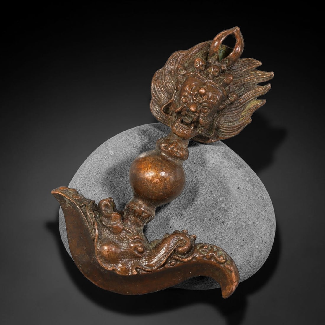 A Tibetan Bronze Buddhist Ritual Implement, 4BP: Cast bronze Tibetan ritual piece, with a curved axe-shaped base featuring mythical creature relief, a bulbous central knop, surmounted by a flaming aureole enclosing a small Buddha figure. A suspensio