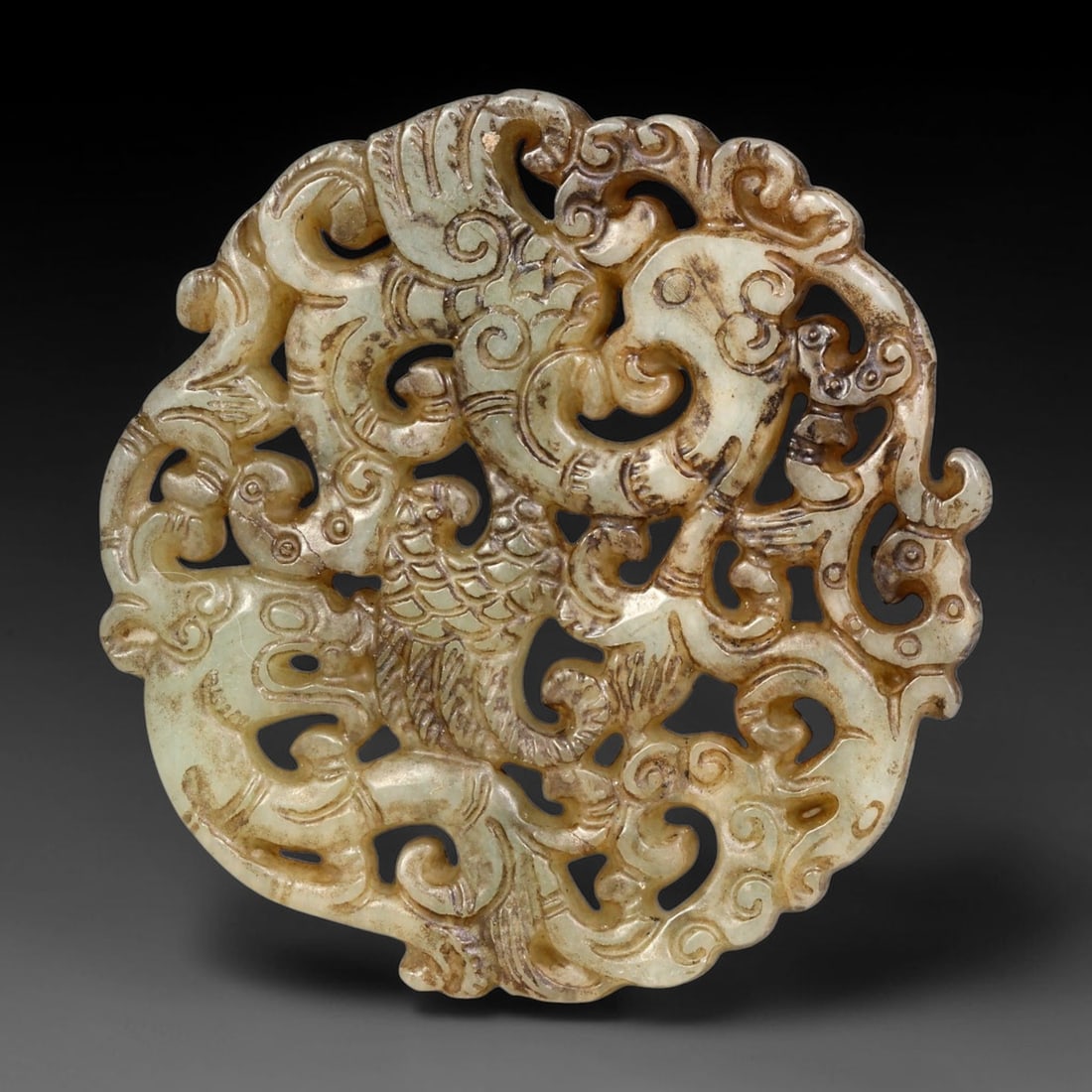 A Chinese Openwork Jade Plaque with Dragon and Phoenix, 82BF: Finely carved pale celadon jade circular plaque with elaborate openwork composition, interweaving dragons and phoenix with scrolling cloud motifs. The stone carries mottled brown inclusions and aged p