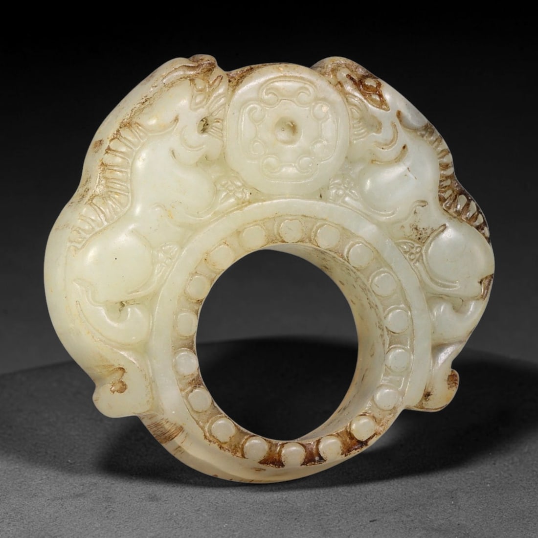 A Chinese Openwork Jade Ring with Twin Mythical Beasts, 84BF: Carved from pale jade with localized brown surface staining along relief edges, the object features a thick annular core decorated with repeating circular bosses on the outer rim. Two mirrored mythica