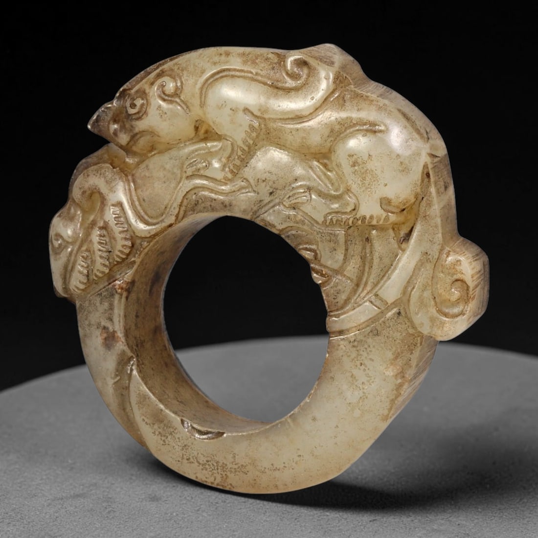 A Chinese Jade Thumb Ring with Carved Chilong Relief, 80BF: A carved jade archer's thumb ring (she ban) of pale celadon tone, intricately decorated in high relief with a coiled chilong dragon crawling over the ring's surface. The stone bears areas of russet an